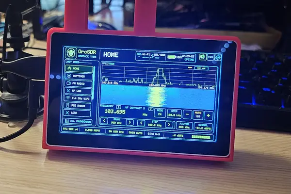 An M5Stack Tab5 on my desk running OrcSDR's Home screen: an FM spectrum and waterfall around 103.7 MHz with tuning controls below