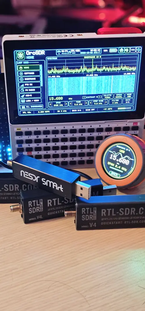 An M5Stack Tab5 with its keyboard running OrcSDR at 15.680 MHz and an M5Stack Dial showing 15.680, with a Nooelec NESDR SMArt, an RTL-SDR Blog V4L and a V4 in front