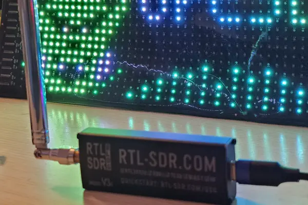 An RTL-SDR Blog V3c with a telescopic antenna and a USB-C cable, in front of the glowing OrcSDR LED sign
